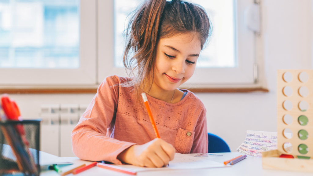 Uma menina jovem está sentada em uma mesa, desenhando com um lápis. Ela sorri levemente enquanto se concentra em seu trabalho. Ao redor dela, há vários materiais de estudo e jogos e lápis coloridos à esquerda. A cena ocorre em um ambiente bem iluminado, sugerindo que ela está em casa ou em um espaço de estudo confortável.